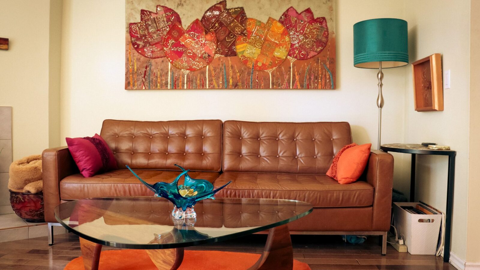 Cozy living room featuring a brown leather sofa, colorful artwork, glass table, and vibrant decor.